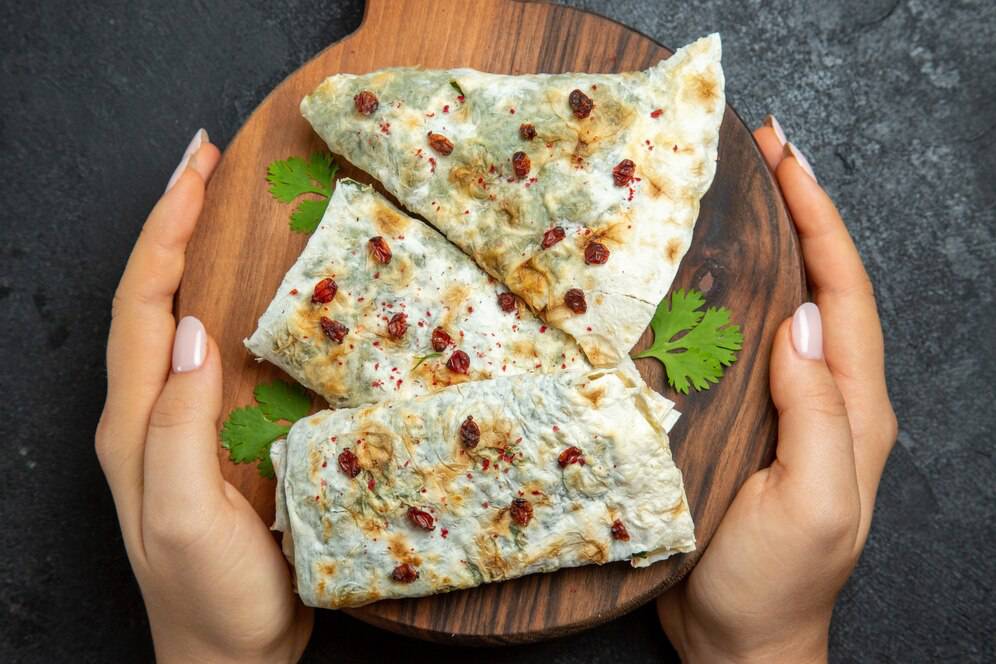Hands holding a wooden board with pieces of garnished flatbread, sprinkled with red spices and herbs, on a dark table.