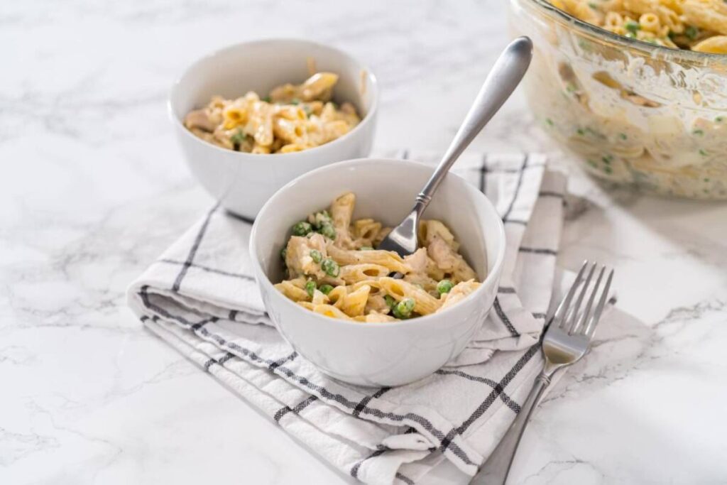 Two bowls of creamy chicken pasta with peas on a marble countertop, one with a fork beside it.