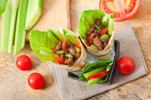 Two beef and vegetable wraps in glass jars on a wooden table, with fresh tomatoes and celery nearby.