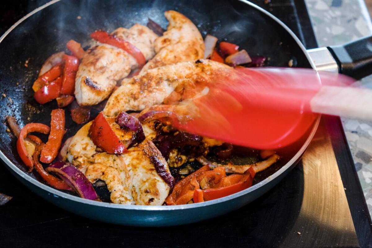 Chicken and bell peppers being stir-fried in a skillet with a red spatula.