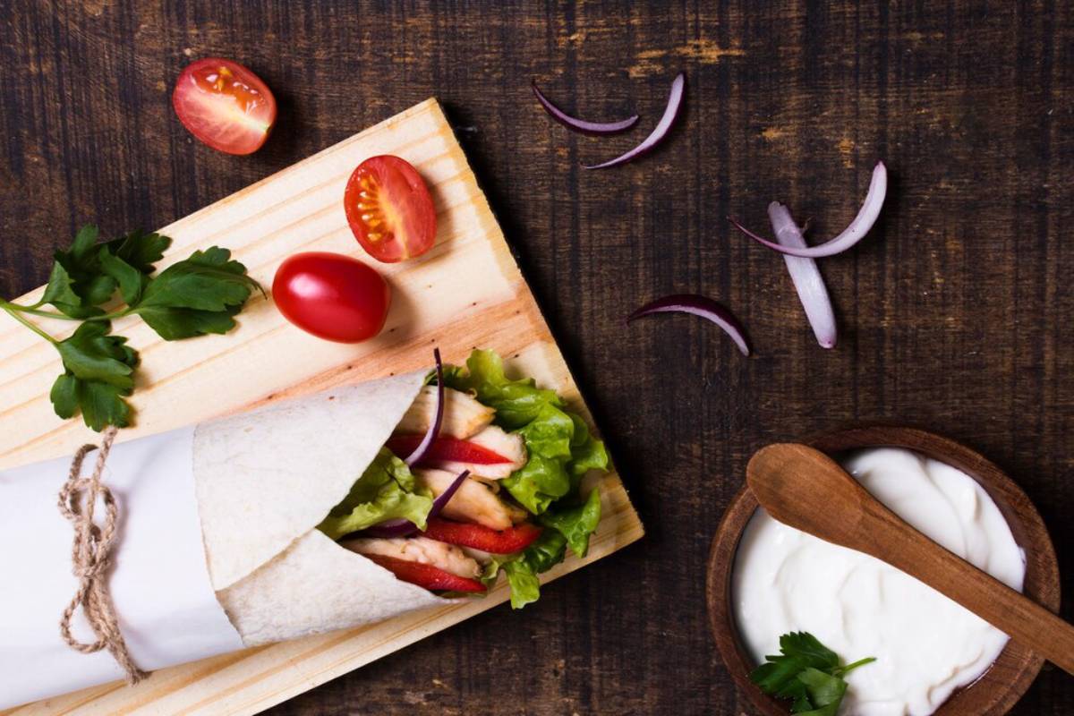 Chicken wrap on a cutting board with tomatoes, onions, and a bowl of yogurt sauce on a wooden table.