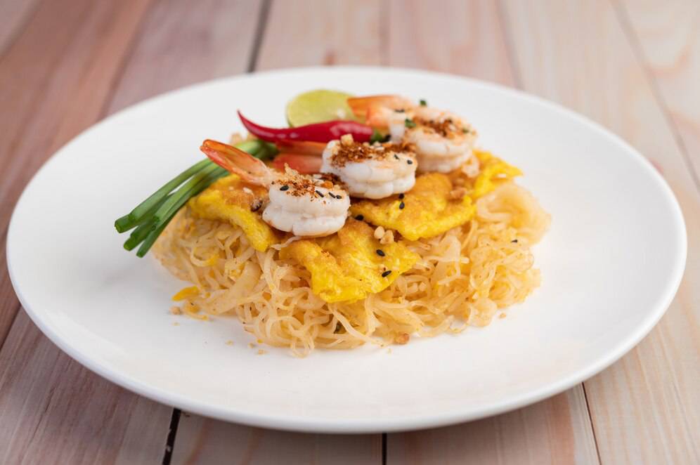 White plate with Thai stir-fried noodles, shrimp, omelet strips, and garnishes on a wooden table.