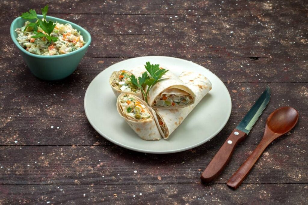 Plate with sliced wraps and a side of coleslaw, with cutlery on a wooden table.