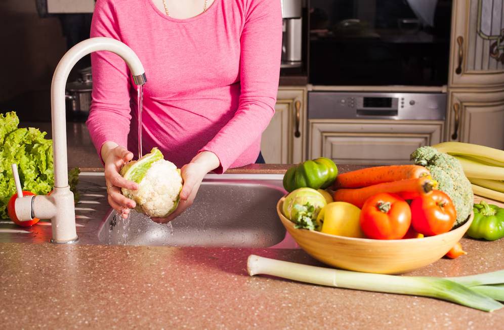 Woman washing cauliflower at kitchen sink, with assorted fresh vegetables nearby.