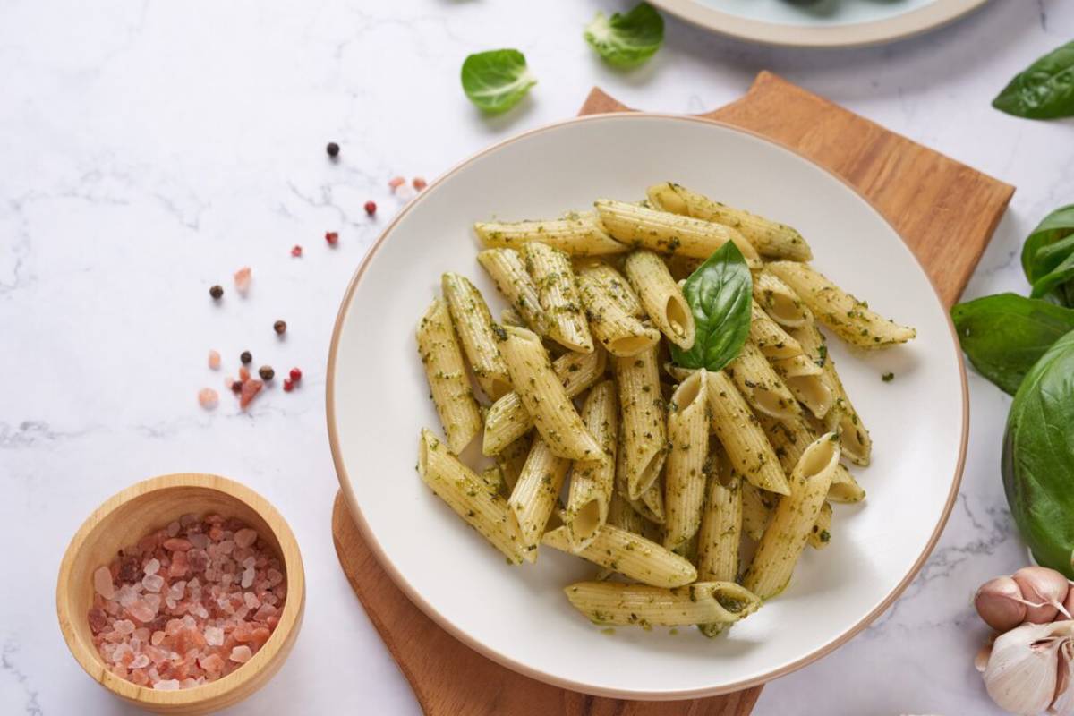 Plate of basil pesto penne pasta, garnished with fresh basil, surrounded by ingredients on a marble countertop.
