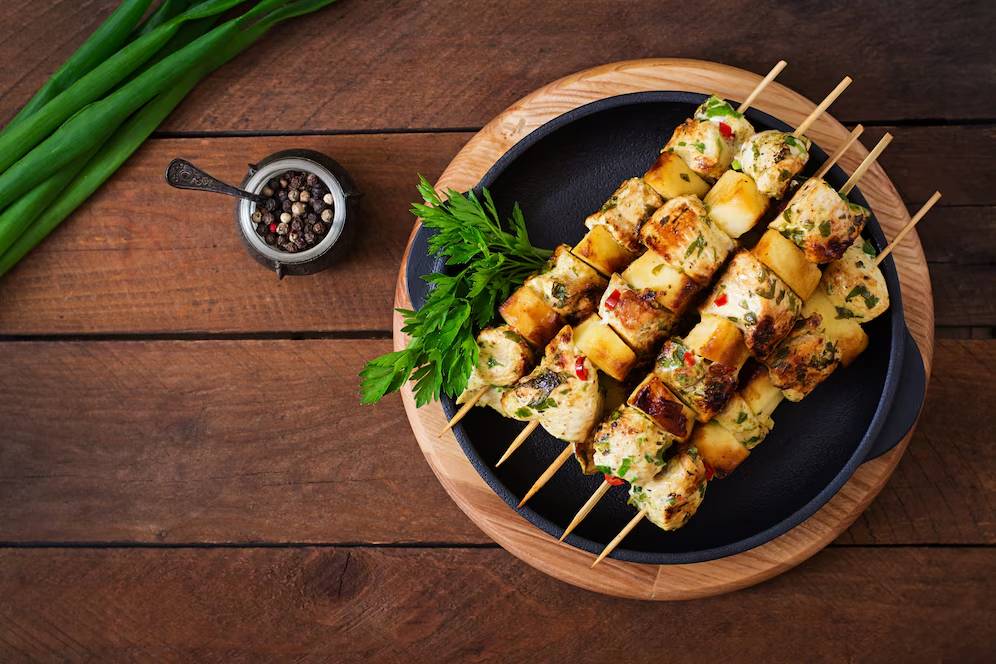 Grilled halloumi cheese skewers, garnished with herbs, served on a black plate with green onions and peppercorns beside it.