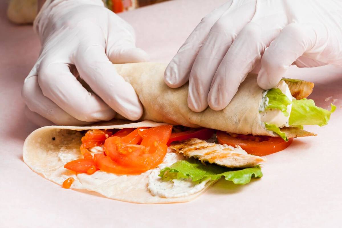 Hands in gloves preparing a chicken wrap with tomatoes, lettuce, and sauce on a flatbread.
