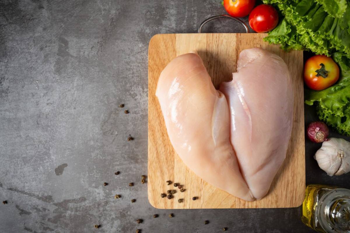 Raw chicken breasts on a wooden cutting board surrounded by fresh vegetables and spices on a grey surface.