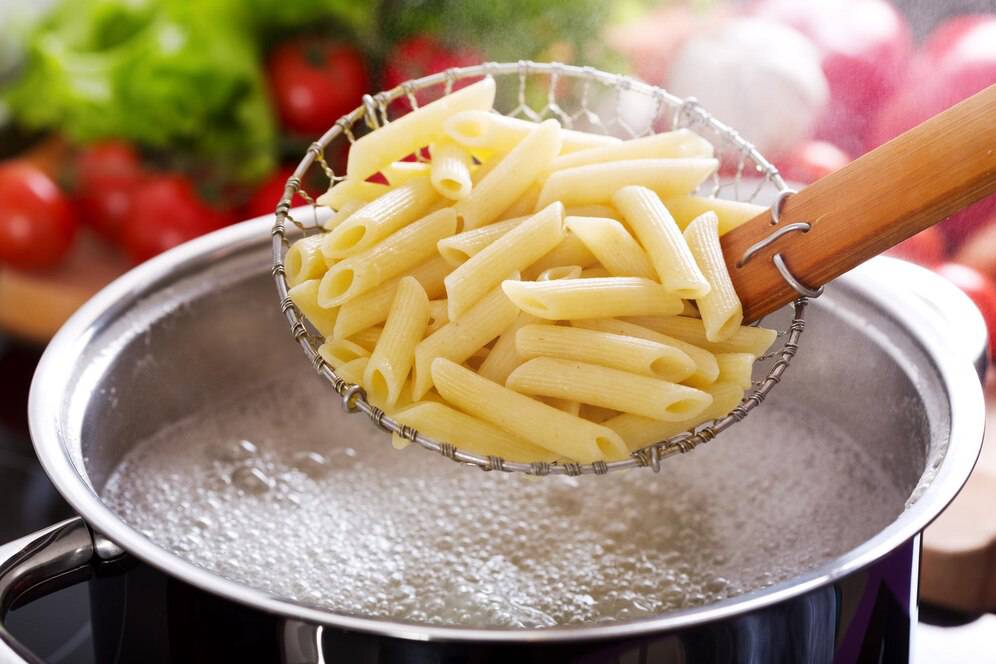 Penne pasta being lifted in a strainer above a boiling pot, with vegetables in the background.