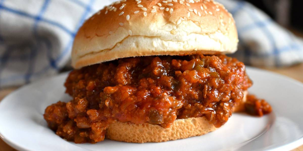 A sesame seed bun filled with a hearty, tomato-based meat mixture, served on a white plate. 