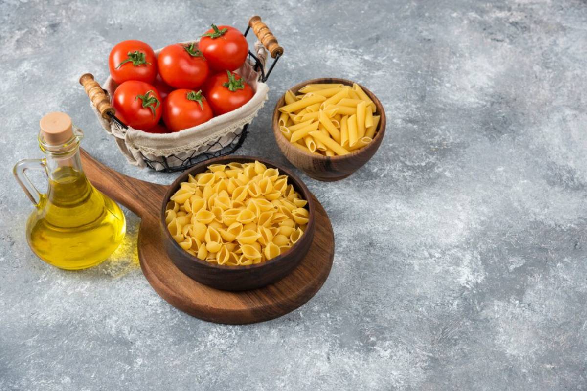 Various pasta types in wooden bowls with tomatoes and olive oil on a textured surface.
