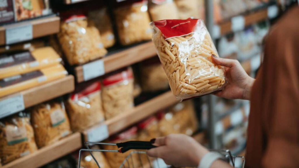 A shopper holds a bag of pasta in front of a shelf stocked with various pasta types in a grocery store.