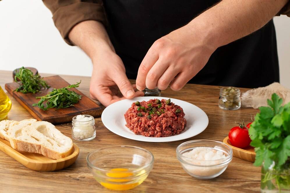 A person garnishing a plate of steak tartare with herbs, surrounded by ingredients like bread, tomatoes, and oil.