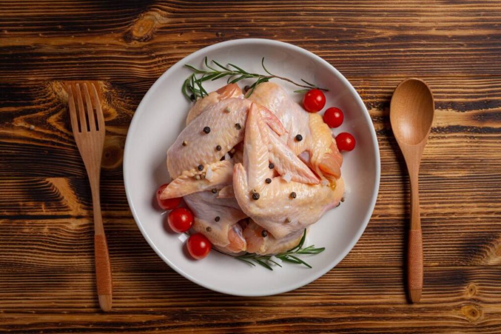 A white plate holds seasoned chicken wings with cherry tomatoes and rosemary, alongside wooden utensils on a rustic wooden table.