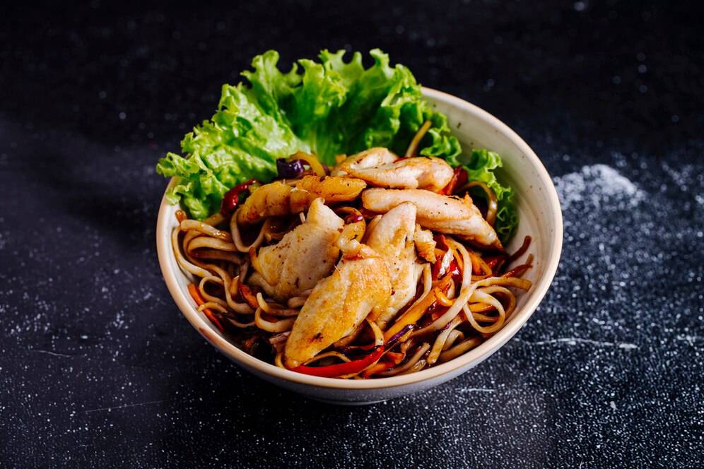Bowl of Asian noodles with sautéed chicken and fresh lettuce on a dark surface.