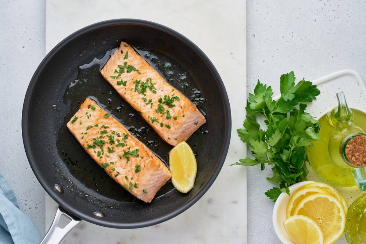 Two salmon fillets seasoned with herbs in a frying pan, garnished with a lemon wedge and parsley, with olive oil nearby.
