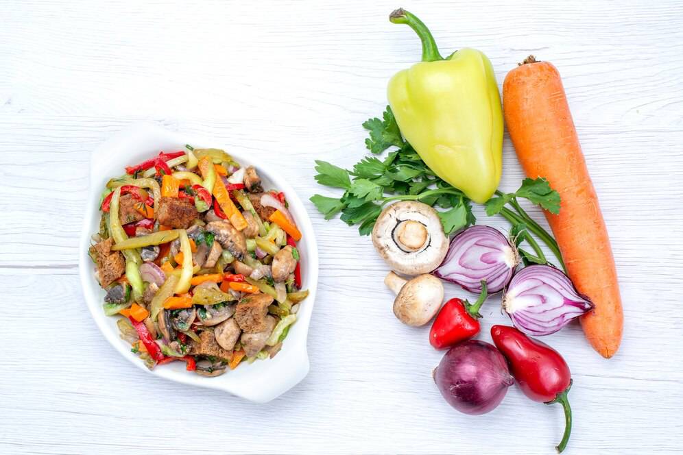 A stir-fried vegetable and meat dish with peppers, onions, carrots, and herbs on a white wooden surface.