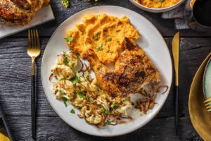 A plate with roasted chicken leg, creamy sweet potato mash, and sautéed cauliflower garnished with herbs on a rustic wooden table.