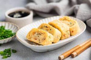 A plate of rolled Japanese omelette garnished with green onion and sesame seeds, alongside soy sauce and chopsticks.