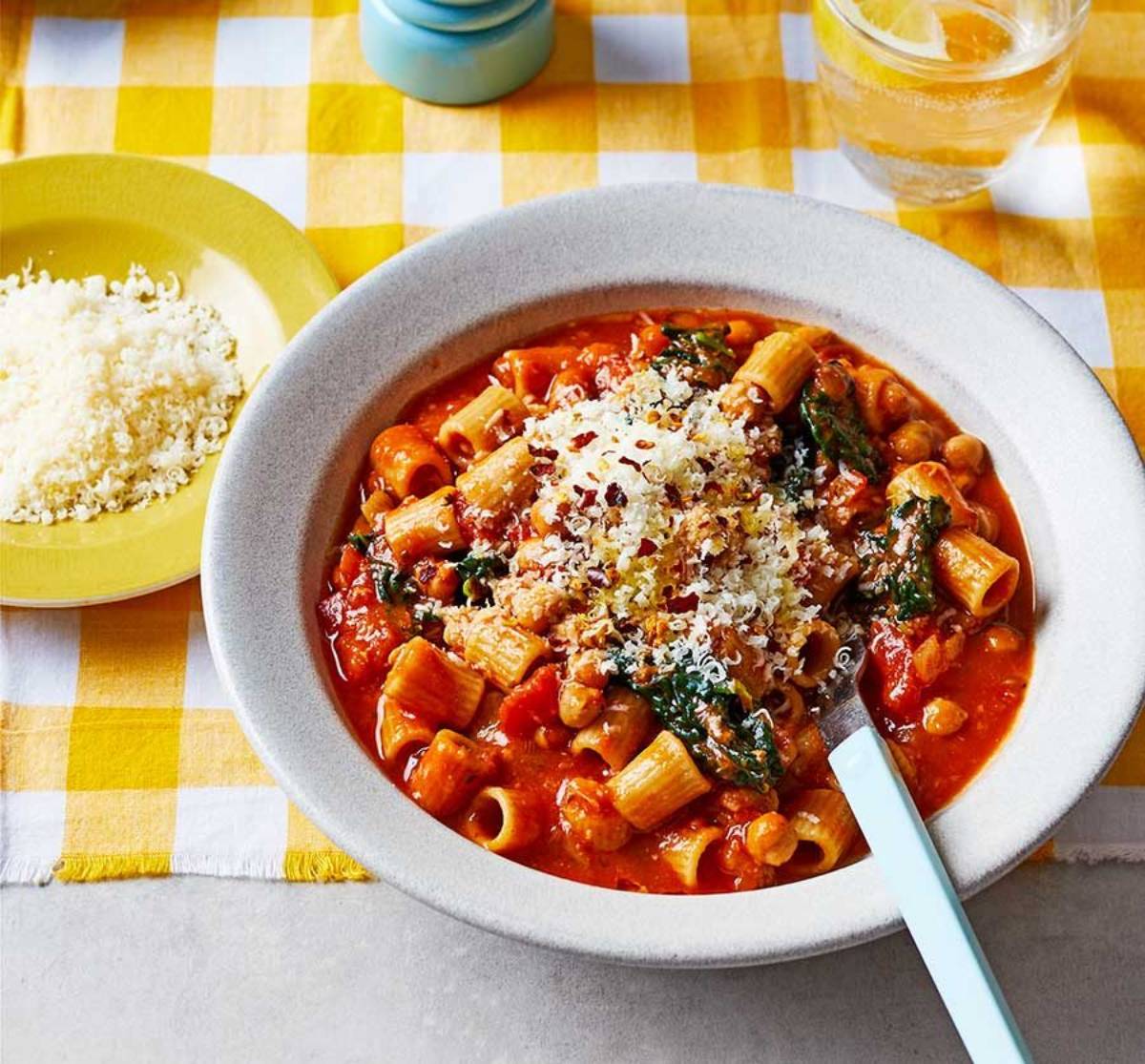 A bowl of pasta with tomato sauce, chickpeas, spinach, and grated cheese, accompanied by a side of couscous.