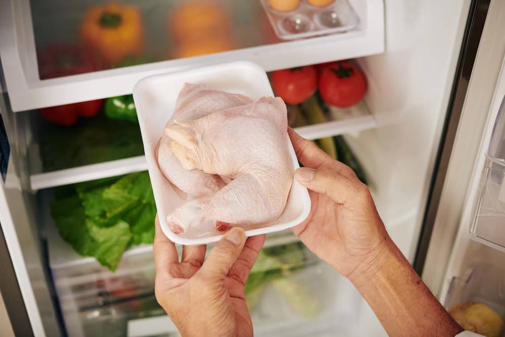 Hands placing fresh chicken in a refrigerator filled with vegetables.