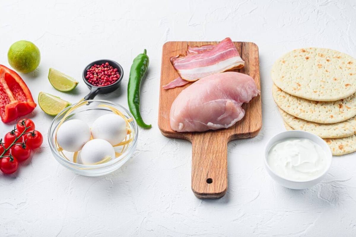 Various fresh ingredients displayed on a white surface: chicken, bacon, eggs, vegetables, lime, and spices.