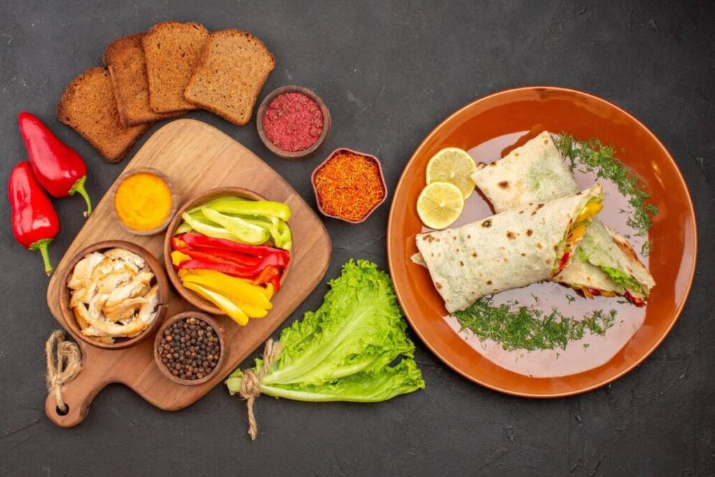 A colorful meal display featuring wraps with veggies, grilled chicken, spices, bread, and fresh lettuce on a dark background.