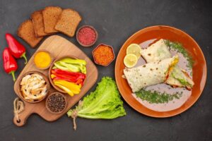 A colorful meal display featuring wraps with veggies, grilled chicken, spices, bread, and fresh lettuce on a dark background.